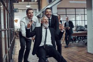 hommes d'affaires joyeux poussant leur patron sur la chaise de bureau tout en courant dans le couloir photo