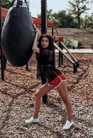 travailler à l'extérieur. belle jeune femme en vêtements de sport regardant la caméra tout en s'appuyant sur le sac de boxe à l'extérieur photo