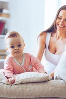 mignon et adorable. petite fille regardant la caméra et s'appuyant sur le coussin pendant que sa mère est assise sur le canapé à l'arrière-plan photo