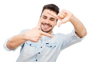 sourire espiègle. beau jeune homme en chemise regardant la caméra à travers le cadre du doigt en se tenant debout sur fond blanc photo