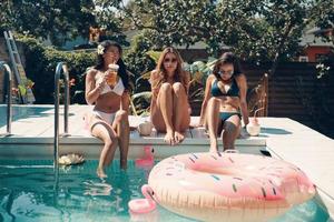 jolies jeunes femmes en bikini buvant des cocktails et souriant assis au bord de la piscine en plein air photo