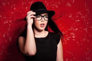 modèle de mode glamour. belle jeune femme au chapeau et lunettes posant sur fond rouge photo