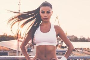 beauté sportive. belle jeune femme en vêtements de sport regardant la caméra et tenant les mains sur la hanche en se tenant debout sur le pont avec vue urbaine en arrière-plan photo