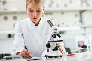 vérification des résultats. jeune femme scientifique sérieuse utilisant un microscope et écrivant dans un bloc-notes assise sur son lieu de travail photo