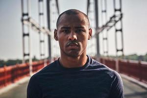 sérieux dans le sport. beau jeune homme africain en vêtements de sport regardant la caméra en se tenant debout sur le pont à l'extérieur photo