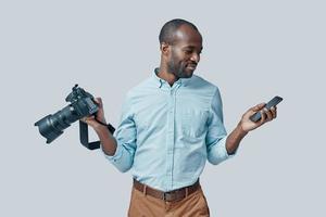 heureux jeune homme africain à l'aide d'un appareil photo numérique et souriant debout sur fond gris