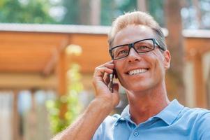 bonne conversation. heureux homme mûr parlant au téléphone portable et souriant tout en étant assis à l'extérieur avec la maison en arrière-plan photo