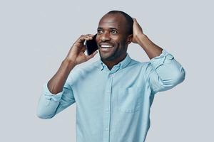 heureux jeune homme africain parlant au téléphone intelligent et souriant debout sur fond gris photo