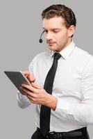 soutenir votre entreprise. beau jeune homme en tenues de soirée et casque travaillant sur tablette numérique en se tenant debout sur fond gris photo