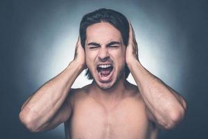 son trop fort. portrait d'un jeune homme torse nu frustré gardant les yeux fermés et couvrant les oreilles avec les mains en se tenant debout sur fond gris photo