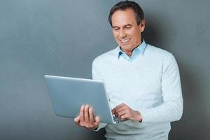 utilisant la technologie pour un grand avantage. joyeux homme mûr travaillant sur ordinateur portable et souriant en se tenant debout sur fond gris photo