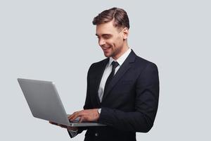 prêt à travailler dur. beau jeune homme en costume complet utilisant un ordinateur portable et souriant debout sur fond gris photo