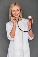 enlevez votre chemise s'il vous plaît belle jeune femme médecin tenant un stéthoscope et regardant la caméra avec le sourire en se tenant debout sur fond gris photo
