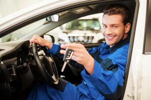 le travail est fait. joyeux jeune homme en uniforme tendant la main avec des clés tout en étant assis dans une voiture à l'atelier photo