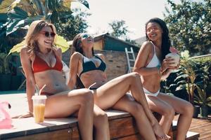 jolies jeunes femmes en maillot de bain buvant des cocktails et souriant assis au bord de la piscine en plein air photo