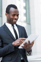 homme d'affaires avec tablette numérique. confiant jeune homme africain en tenue de soirée travaillant sur une tablette numérique et souriant tout en se tenant à l'extérieur photo