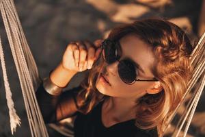 se sentir dragueur. vue de dessus d'une jeune femme attirante ajustant ses lunettes tout en étant assise dans un hamac sur la plage photo