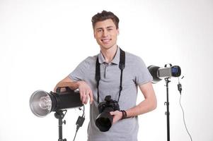 prêt pour le tir. beau jeune homme en polo tenant un appareil photo numérique et souriant tout en se tenant en studio avec un équipement d'éclairage sur fond