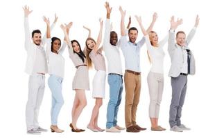 habitué au succès. toute la longueur du groupe de jeunes heureux en tenue décontractée intelligente debout dans une rangée et gardant les bras levés en se tenant debout sur fond blanc photo