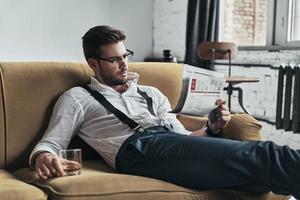 article intéressant. jeune homme élégamment habillé lisant un journal et tenant un verre assis sur un canapé