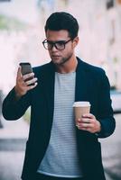 en tapant un message rapide à un ami. beau jeune homme en tenue décontractée intelligente tenant une tasse de café et regardant son téléphone intelligent tout en marchant le long de la rue photo