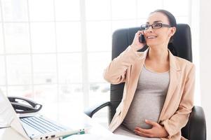 femme d'affaires enceinte au travail. joyeuse femme d'affaires enceinte parlant au téléphone alors qu'elle était assise à son lieu de travail au bureau photo