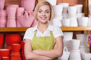 vendre les pots de fleurs. belle jeune femme aux cheveux blonds en tablier gardant les bras croisés debout contre l'étagère avec des pots de fleurs photo