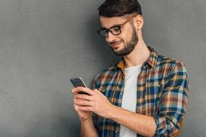 message à elle. joyeux jeune homme dans des verres à l'aide de son smartphone avec le sourire en se tenant debout sur fond gris photo