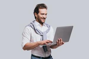 obtenir les meilleurs résultats. beau jeune homme en vêtements décontractés intelligents travaillant sur l'ordinateur portable et souriant en se tenant debout sur fond gris photo