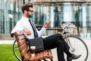 jeune et prospère. vue latérale d'un beau jeune homme d'affaires tenant un téléphone portable assis sur le banc près de son vélo avec un immeuble de bureaux en arrière-plan photo