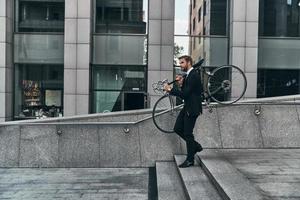 en évitant le trafic. toute la longueur du jeune homme en costume complet portant son vélo tout en marchant à l'extérieur photo