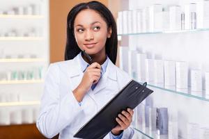 penser à un médicament approprié pour vous. jeune femme africaine réfléchie en blouse de laboratoire tenant le presse-papiers et regardant loin avec le sourire tout en se tenant près de l'étagère de la pharmacie photo