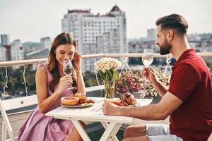 jeune couple insouciant dans des vêtements décontractés appréciant un dîner romantique et souriant tout en étant assis sur la terrasse sur le toit à l'extérieur photo