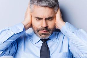 se sentir stressé et surmené. homme mûr frustré en chemise et cravate couvrant les oreilles avec les mains et gardant les yeux fermés en se tenant debout sur fond gris photo