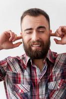 entendre aucun mal. beau jeune homme barbu faisant une grimace et couvrant les oreilles par les doigts en se tenant debout isolé sur fond gris photo