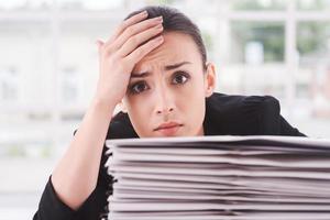 fatigué et surmené. jeune femme déprimée en costume regardant par la pile de documents posés sur la table et touchant son front avec la main photo