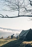 il est l'allume-feu. jeune homme faisant un feu de camp assis près de la tente dans les montagnes photo