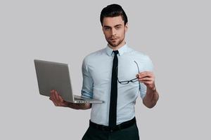 expert en affaires confiant. beau jeune homme en chemise blanche et cravate tenant un ordinateur portable et des lunettes en se tenant debout sur fond gris photo