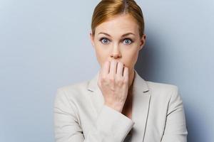 un peu nerveux. jeune femme d'affaires nerveuse se mordant les ongles en se tenant debout sur fond gris photo