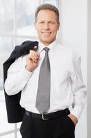 Un homme d'affaires qui réussit. joyeux homme mûr en chemise et cravate tenant la veste par son doigt et souriant à la caméra tout en se tenant près de la fenêtre photo