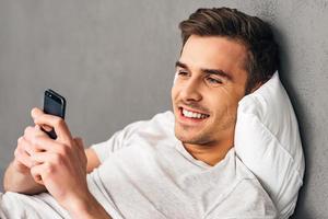 première chose que vous faites après le réveil. joyeux jeune homme tenant un téléphone intelligent et le regardant avec le sourire en position couchée sur un oreiller sur fond gris photo