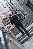 prendre un vélo. vue arrière sur toute la longueur du jeune homme en costume complet portant son vélo tout en marchant à l'extérieur photo