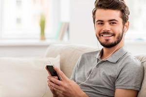 appréciant sa musique préférée. joyeux jeune homme dans un casque tenant un lecteur mp3 et regardant la caméra tout en étant assis sur un canapé photo