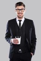 prendre le temps d'une pause-café. confiant jeune homme charmant en tenues de soirée et lunettes tenant une tasse de café et souriant en se tenant debout sur fond gris photo