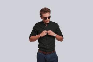 style parfait. beau jeune homme à lunettes boutonnant sa chemise en se tenant debout sur fond gris photo