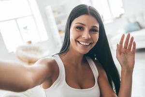 prendre un joli selfie. autoportrait de belle jeune femme regardant la caméra et souriant assis sur le canapé à la maison photo