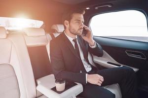 entreprise en mouvement. beau jeune homme en costume complet parlant au téléphone intelligent alors qu'il était assis dans la voiture photo