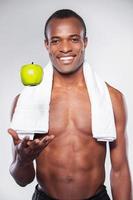 profiter d'un mode de vie sain. jeune homme africain musclé avec une serviette sur l'épaule vomir une pomme et souriant à la caméra en se tenant debout sur fond gris photo
