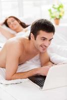 surfer sur le web au lit. joyeux jeune homme utilisant un ordinateur portable en position couchée dans son lit avec sa petite amie photo