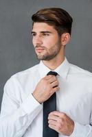 jeune et prospère. confiant jeune homme d'affaires ajustant sa cravate et regardant ailleurs en se tenant debout sur fond gris photo